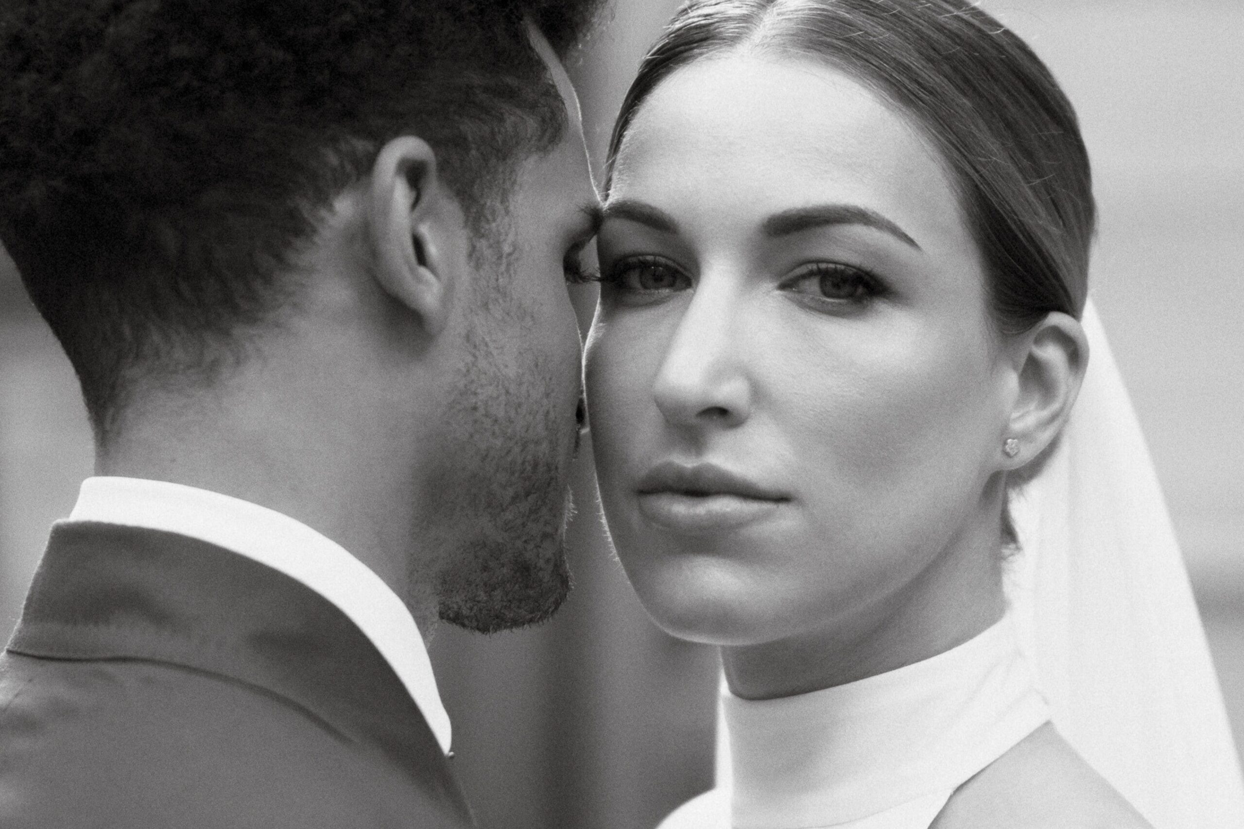 Black and white portrait of bride and groom, with bride facing the camera and the groom facing away, both leaning in to each other.