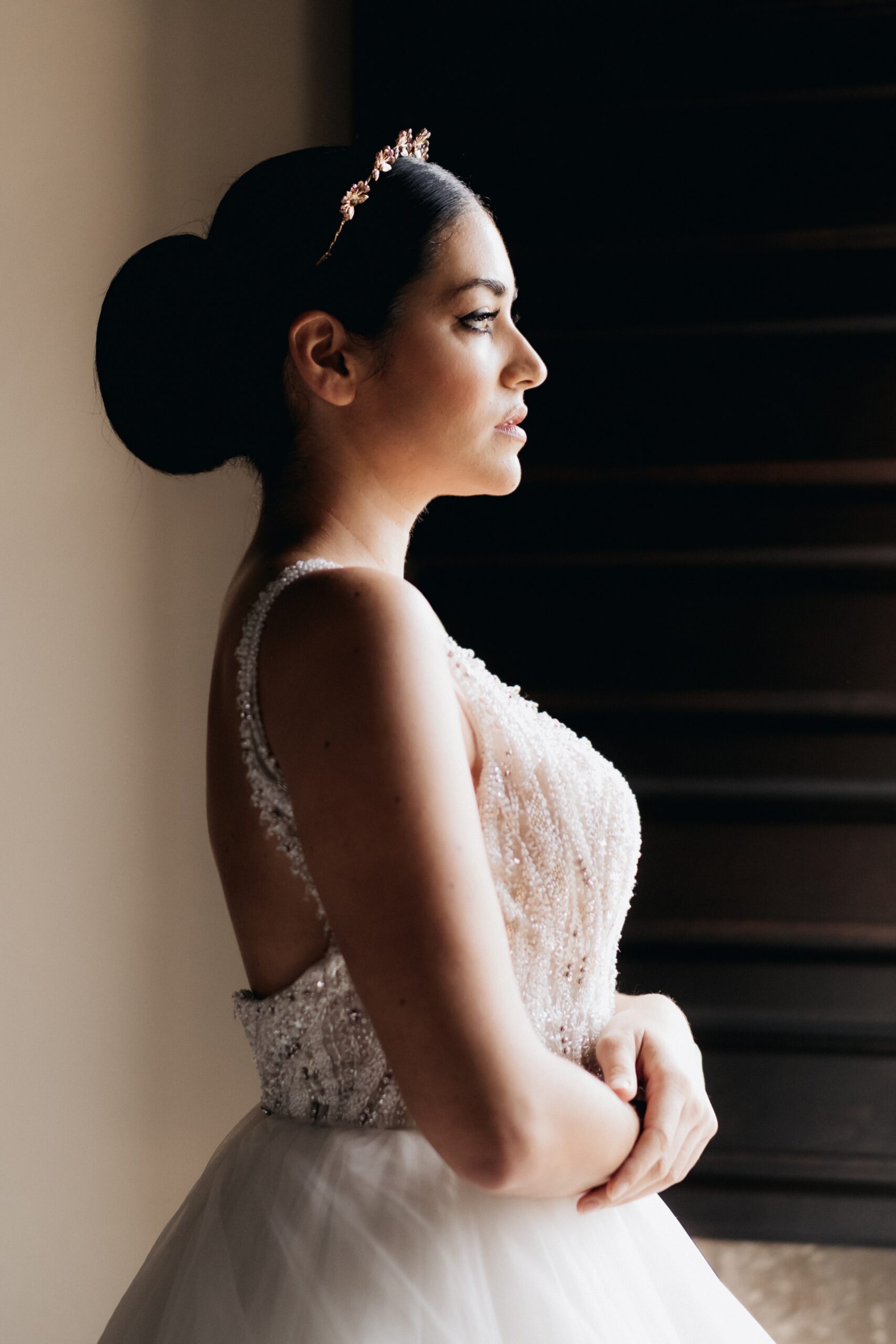 Profile photo of bride wearing a white grown and golden crown with hair in a slick bun facing a window and crossing her arms.