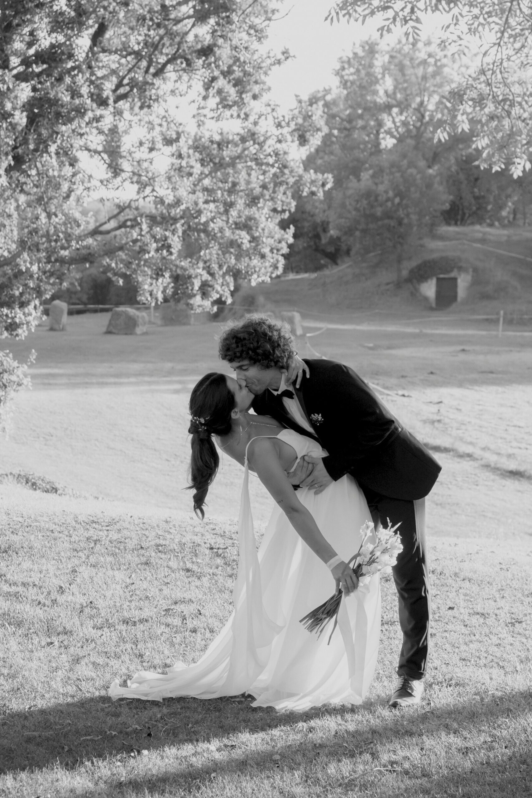 Black and white photo of groom leaning bride backwards and kissing her on their wedding day in front of a field under a tree.